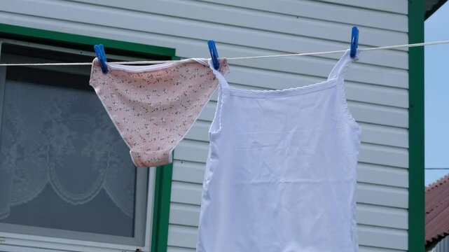 A pair of pink cotton panties and a white cotton camisole are hung on a clothesline under a clear sky. This image reflects the natural simplicity of drying clothes outdoors, blending comfort and