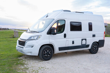 Front and side of a modern white camper van with roof cooler, camped in an empty camper area in the wilderness at sunset.