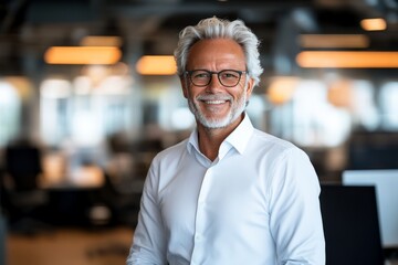 Fototapeta premium A mature man with stylish gray hair and glasses smiles warmly while standing in a modern office, exuding wisdom, professionalism, and a friendly demeanor.