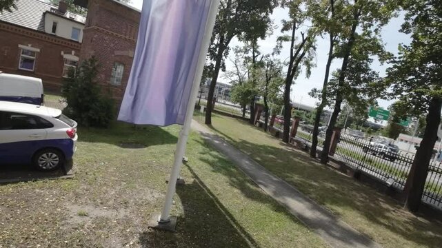 A collection of flags on flagpoles are waving in the wind in front of a building . There are five flagpoles. On the left is a yellow flag with a blue stripe down the center and a coat of arms on the