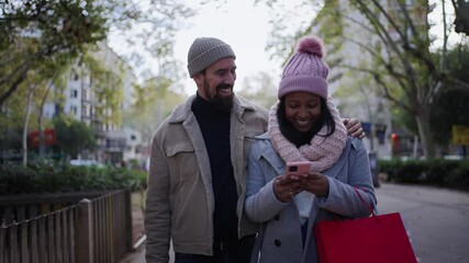 Beautiful young couple interracial in love using mobile phone enjoying winter holiday season in the city streets, walking and hugging and holding gift bags while celebrating Christmas outdoors