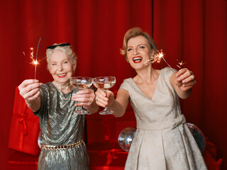 Two beautiful stylish mature senior women in dresses with sparklers and glasses celebrating new...