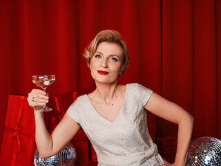 mature stylish elegant woman in cocktail dress with glass of sparkling wine with presents on red background. Happy New Year!