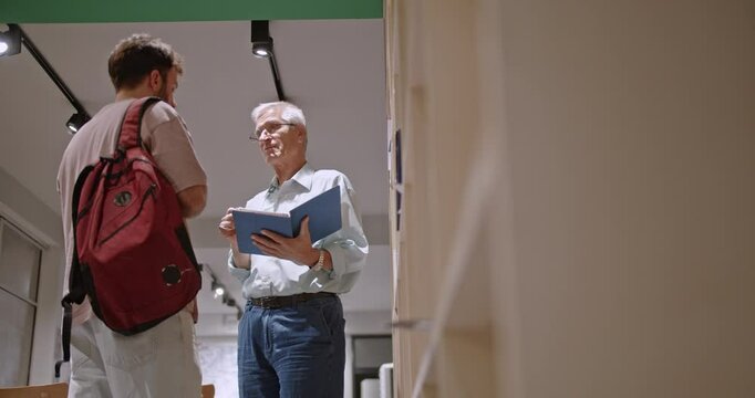 A senior professor explains the homework assignment to a young male student at the university library, offering guidance and clarification as the student takes notes attentively.
