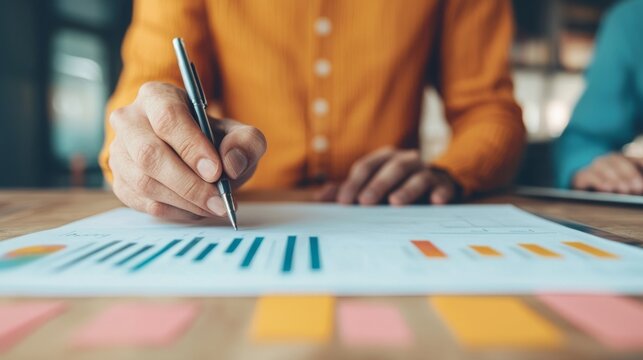 A person analyzes graphs and data on paper while writing with a pen, showcasing a collaborative work environment in a modern office.