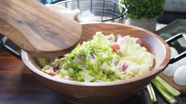 Making potato salad with potatoes, onion, sauce, celery and mixing it around in a wooden bowl. Cold side dish for family gatherings and cook outs.