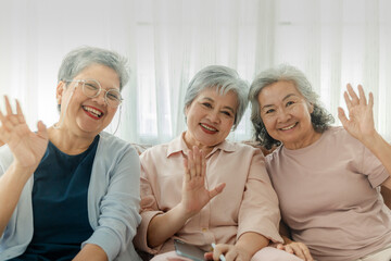 Three elderly women sit together, smiling and waving at the camera, radiating joy and friendship. The image emphasizes companionship, happiness, and togetherness in a relaxed, homey setting.