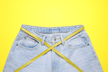 Jeans and measuring tape on yellow background, top view