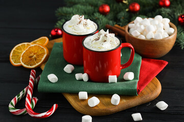 Tasty hot cocoa drinks with whipped cream in mugs, marshmallows and candy canes on black wooden table, closeup
