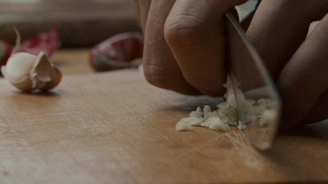 Close-up view of hands finely chopping garlic on wooden cutting board with some unpeeled garlic cloves in the background