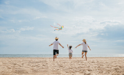 Asian family is flying a kite high in the sky while running on the beach. Happy parents mother and father with their child playing with kite, family is enjoying their time together, summer day