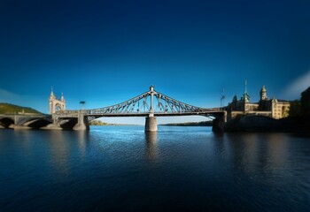 Obraz premium A metal bridge with a truss design crosses a river against a clear blue sky, buildings and trees line the riverbanks.