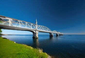 Naklejka premium A white metal bridge with a small tower spanning a body of water with a blue sky above.