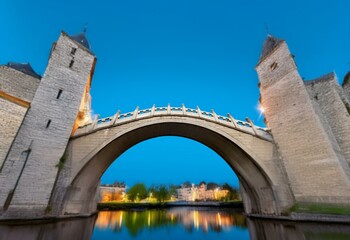 A stone arch bridge spans a calm river, with two stone towers rising on either side against a clear blue sky.