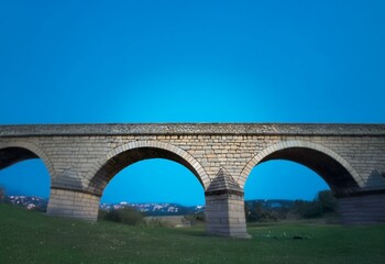 Fototapeta premium Stone arch bridge with a blue sky background, grass in the foreground.