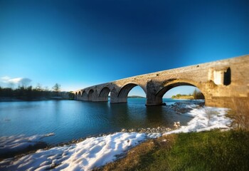 Fototapeta premium Stone arch bridge over a blue water body, with white snow on the banks.