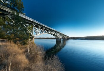 Obraz premium A steel bridge arches over a calm river with a deep blue sky overhead.