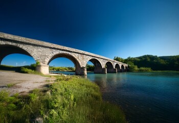 Fototapeta premium A stone bridge arches over a river, with lush green vegetation bordering the waterway.
