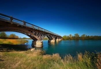 Fototapeta premium A wooden bridge with a stone foundation spanning a river under a clear blue sky.