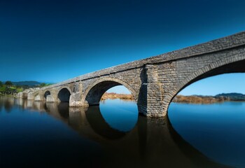 An ancient stone bridge arches over calm blue water, its reflection mirroring the structure below.
