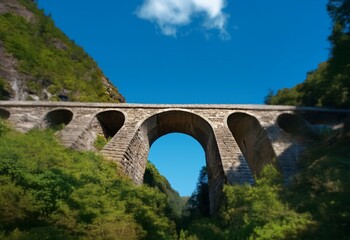 Fototapeta premium A stone arch bridge spans a valley, framed by green trees and a bright blue sky with a single fluffy cloud.