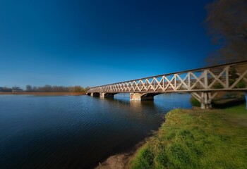 Fototapeta premium A wooden bridge spans a river with calm water against a bright blue sky.