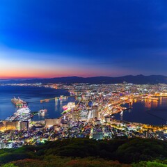 Fototapeta premium 函館山から見下ろす、世界三大夜景のひとつとされる函館の夜景。湾に沿って広がる街の光が宝石のように輝き、港に停泊する船の灯りも穏やかに瞬いている。ロマンチックな夜景を眺めるカップルや観光