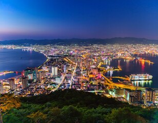 Fototapeta premium 函館山から見下ろす、世界三大夜景のひとつとされる函館の夜景。湾に沿って広がる街の光が宝石のように輝き、港に停泊する船の灯りも穏やかに瞬いている。ロマンチックな夜景を眺めるカップルや観光