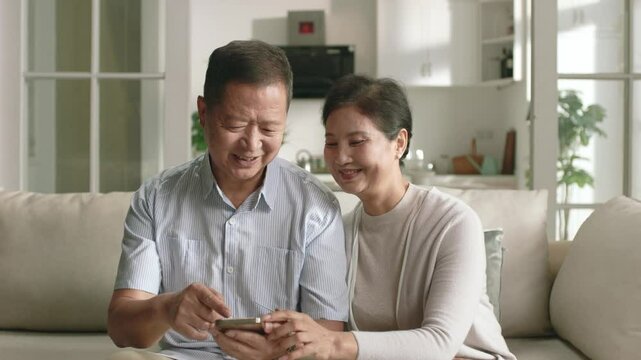 happy loving senior asian couple sitting on family couch looking at cellphone together