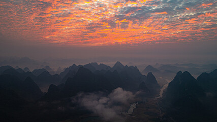 .A breathtaking aerial view of a dramatic sunrise illuminating the sky with vibrant orange and red hues over mist-covered karst mountains. The serene landscape, dotted with valleys and rivers,.