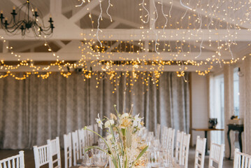 In a spacious room adorned with lights hanging elegantly from the ceiling, there is a long dining table beautifully decorated with a vase of vibrant flowers