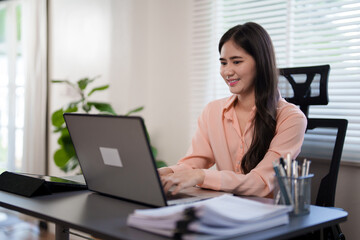 A young woman is sitting at her desk in an office, working on her laptop. She looks focused and engaged, with office documents and a coffee mug beside her, in a bright, modern workspace