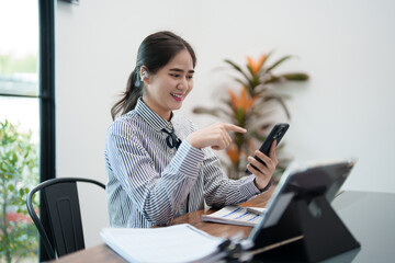 A young woman is smiling and looking at her phone while sitting at her desk. She appears cheerful and relaxed, wearing headphones in a bright, modern office environment.