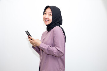 Portrait of emotional Asian, Indonesian woman wearing purple shirt and hijab headscarf laughing and holding handphone. Isolated on white background