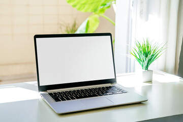 Fototapeta premium Laptop on a desk with white screen for mockup. There are no people on the scene. Computer next to the window. Stock photographs with clear, bright colors.
