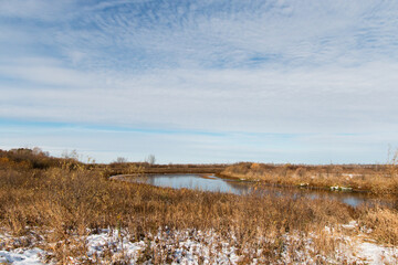Autumn landscape. Cold, blue sky above the horizon. Sunny day. First, white snow melts on a field with dry grass. There is a lake in the background.