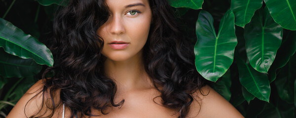 Brunette woman with voluminous curly hair and a confident gaze poses against a backdrop of lush tropical green leaves. Ideal for promoting beauty, haircare, fashion, or nature-inspired products.