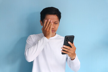 Young Asian man looking at his phone with a sad expression while covering his face