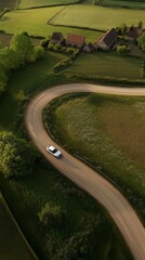 Aerial view of a white car driving along a winding dirt road through lush green countryside with scattered barns.