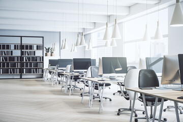 Perspective view on rows of work places in spacious sunny coworking office with modern computers, wooden tables and floor, dark bookcase, white lamps and huge windows with city view. 3D rendering