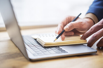 Close-up depiction of a man's hand taking notes in a notepad on a sleek laptop, with a blurred background