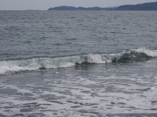 Fototapeta premium 波打ち際の波が崩れる瞬間。日本 小田原市御幸が浜海岸。背景に真鶴半島を望む The moment when the waves break on the beach. Miyukigahama beach, Odawara City, Japan. Manazuru Peninsula in the background.