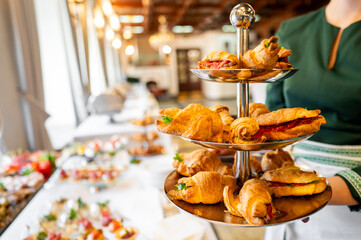A three-tiered serving tray with assorted pastries, held by a person in a green top, with a blurred buffet background. Perfect for high tea, catering, or dessert presentations.