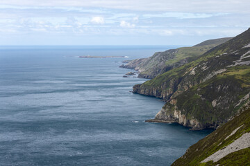 The dramatic cliffs of Slieve League in Ireland stretch along the Atlantic Ocean, with steep, rocky faces and verdant grassland. A small island with a lighthouse is visible offshore