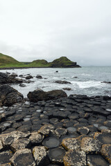 Vertical image of Giant's Causeway in Northern Ireland, highlighting hexagonal basalt columns in the foreground with ocean waves, green hills, and cloudy sky