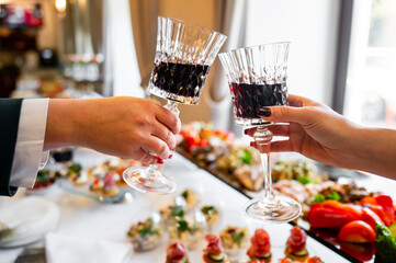 Two hands holding glasses of red wine, toasting over a blurred background of a food buffet. The image captures a celebratory moment, perfect for themes of dining, celebration, and social gatherings.