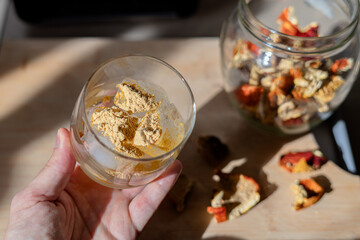 A glass filled with ice cubes and powdered Amanita muscaria mushroom on its surface, held in the hand, illuminated by natural sunlight. Preparing a homemade fly agaric elixir