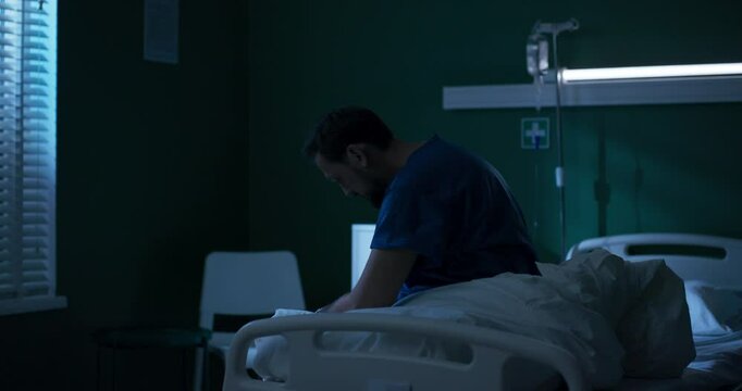 Desperate man seated at edge of bed, trembling, gazes hopelessly out the window, lost in thoughts.