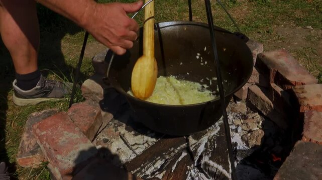 A traditional Romanian cooking scene showing onions being saut&eacute;ed in a cauldron over a wood fire, a key step in making authentic zacusca. The rustic outdoor setup highlights the time-honored methods.