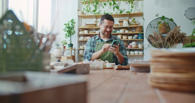 Young Caucasian male with beard and glasses sitting at table in studio and texting on smartphone. Typing message in chat. Reading funny text from friend and laughing. Resting from working.